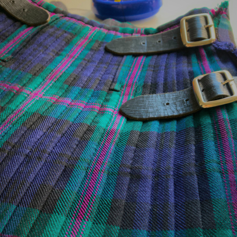 Kilt alteration Greenock A close-up of a tartan kilt with buckles, featuring green, black, and pink patterns.
